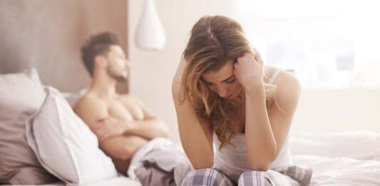 Libido em Baixa (causas, sintomas e como tratar) Mulher triste e preocupada, parceiro ao fundo na cama.