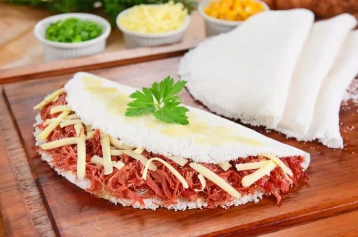 Tapioca recheada com carne e queijo Tapioca recheada com carne, queijo e acompanhamentos em uma tábua de madeira.