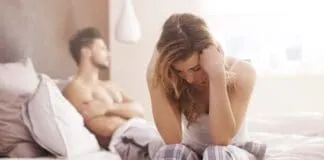 Mulher triste e preocupada, parceiro ao fundo na cama.