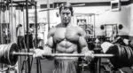 Arnold Schwarzenegger treinando bíceps com barra em academia.