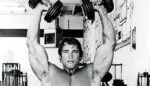 Arnold Schwarzenegger treinando ombros com halteres em academia.