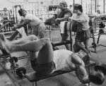 Homem treinando peito com peso na academia, estilo clássico de Arnold Schwarzenegger.