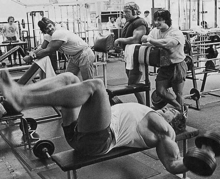 Homem treinando peito com peso na academia, estilo clássico de Arnold Schwarzenegger.