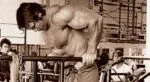 Arnold Schwarzenegger treinando peito com barra em academia.
