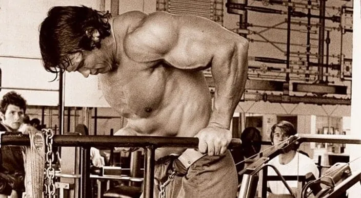 Arnold Schwarzenegger treinando peito com barra em academia.