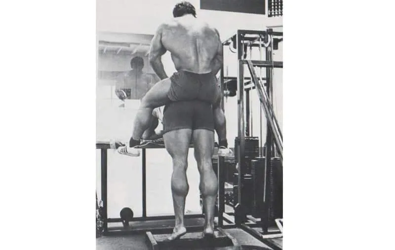 Arnold Schwarzenegger treinando pernas com parceiro no ginásio.