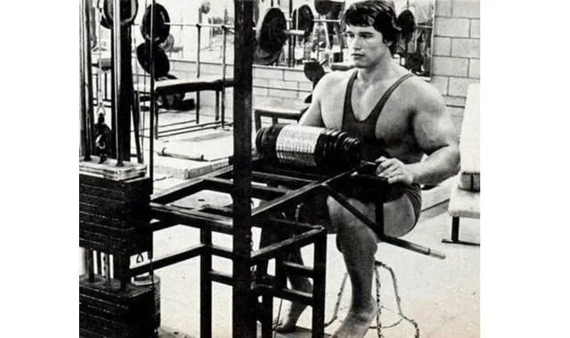 Arnold Schwarzenegger treinando pernas com halteres na academia.