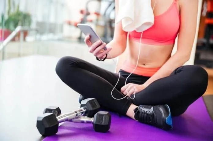 Mulher sentada na academia usando celular com fones de ouvido.